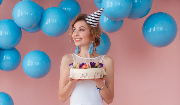 Young woman holding cake with macaroon decor on pink background with blue balls hanging. Full lenght portrait